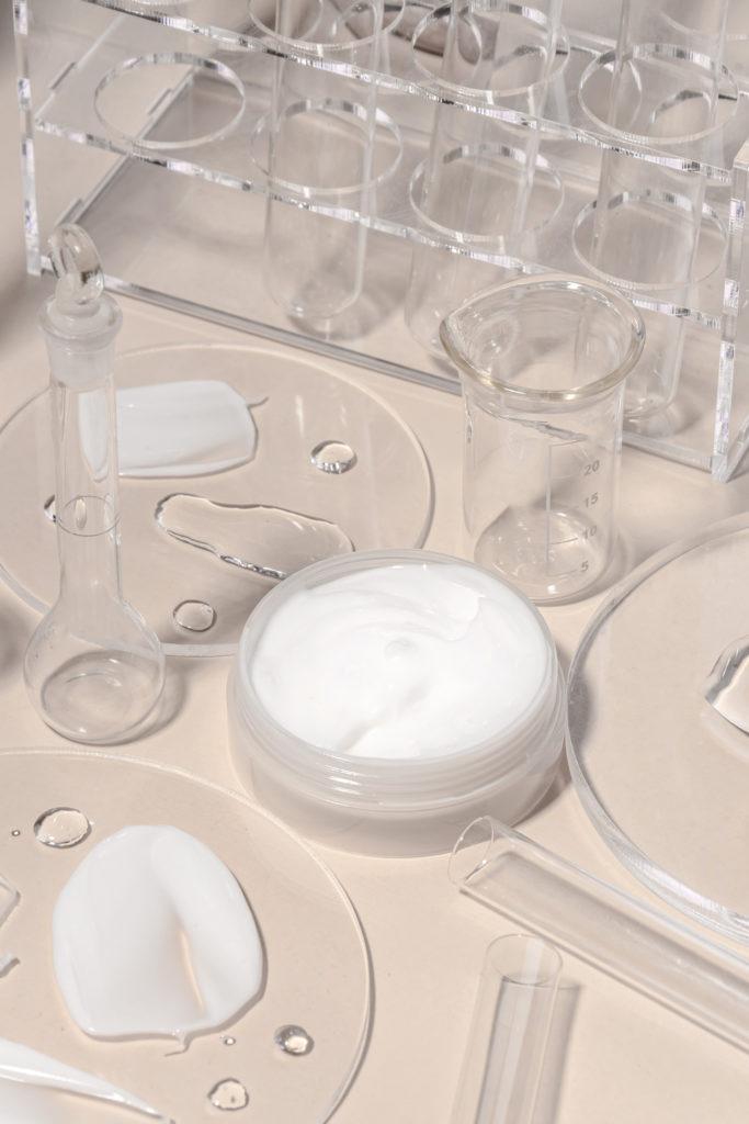 Laborgeräte aus Glas wie Reagenzgläser, Becherglas und Reagenzglasständer neben transparenten Platten mit weißen Crememustern auf hellem Untergrund.