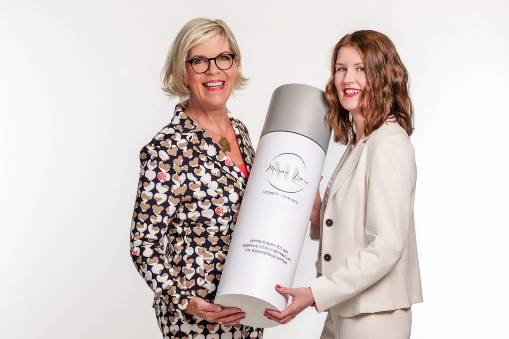 Zwei lächelnde Frauen, eine mit blondem Haar und gemusterter Jacke mit Herzen, die andere mit braunen Haaren und hellbeigem Blazer, halten zusammen eine große, zylinderförmige Dose mit der Aufschrift 'Meine Linie COSMETIC-COMMUNITY Gemeinsam für ein starkes Unternehmertum im Kosmetikgewerbe' vor weißem Hintergrund.