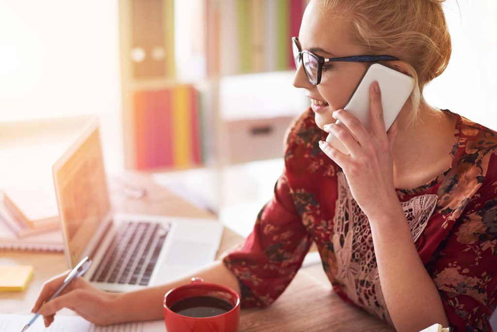 Frau mit Brille und rotem Blumenkleid, die lächelnd am Schreibtisch sitzt, mit einem Smartphone am Ohr telefoniert und mit der anderen Hand Notizen schreibt, vor ihr ein Laptop und eine rote Kaffeetasse.